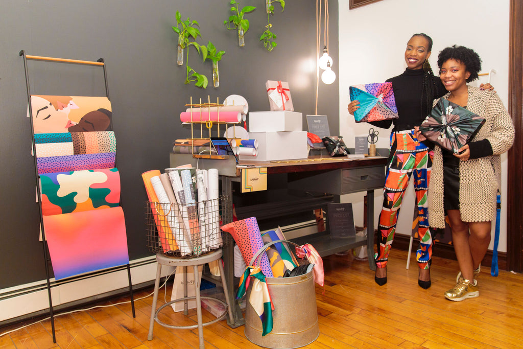 Two women standing and holding boxes wrapped in colorful fabric in home design studio