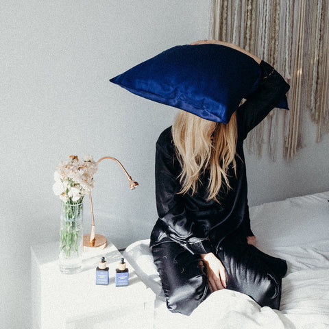 girl in silk pajamas sitting in bed holding silk pillow on top of her head