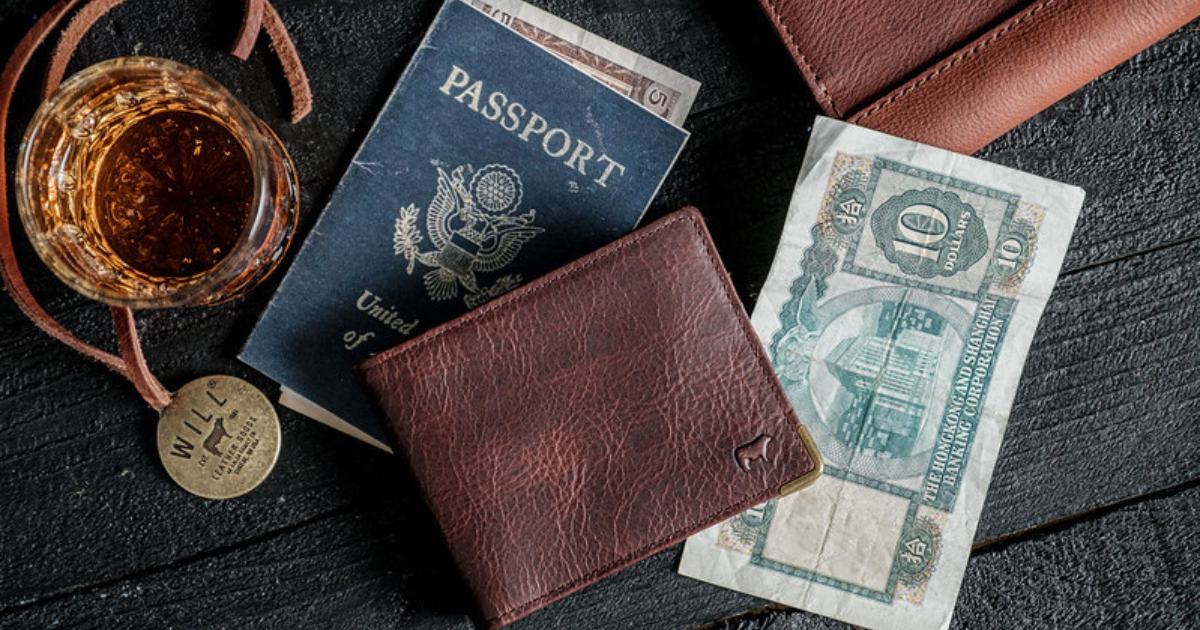 A brown leather walled by Will Leather Goods next to a passport, money, and drink on a table.