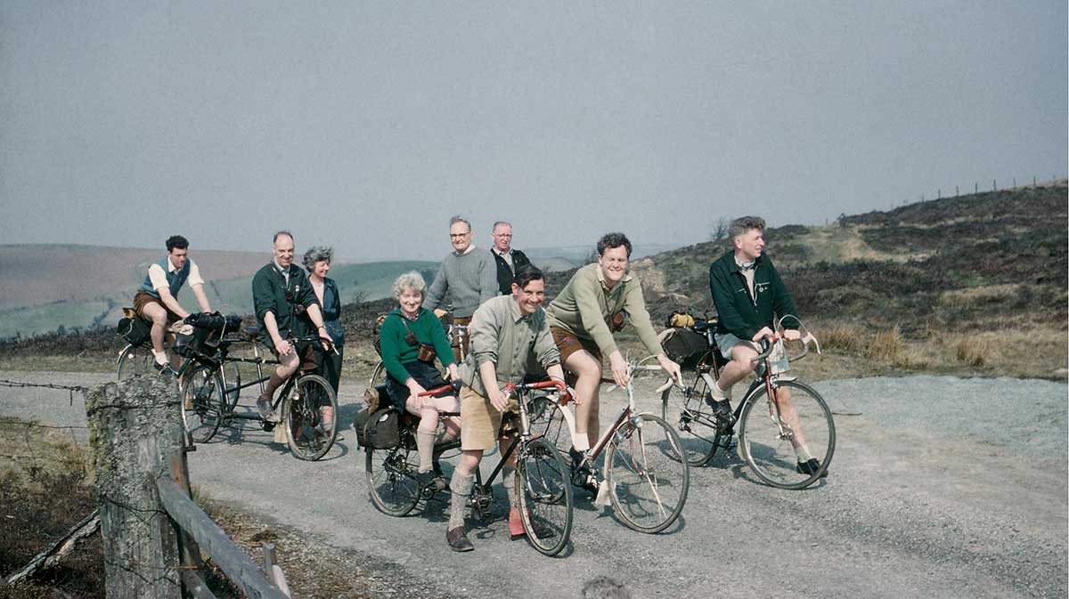 A group of Rough Stuff Fellowship members with their bicycles on a cycling trip in the countryside with fields, forests and mountains.