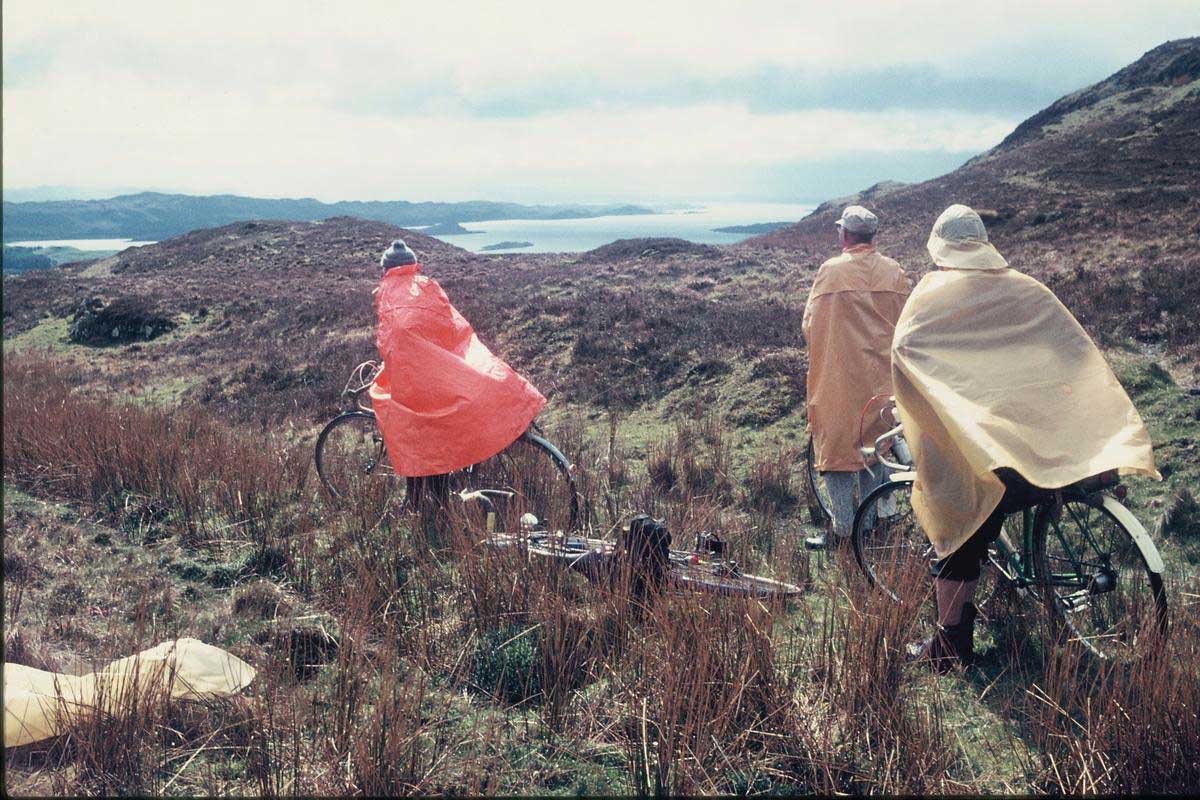 A group of Rough Stuff Fellowship members with their bicycles on a cycling trip in the countryside with plastic ponchos on and a lake in the background.