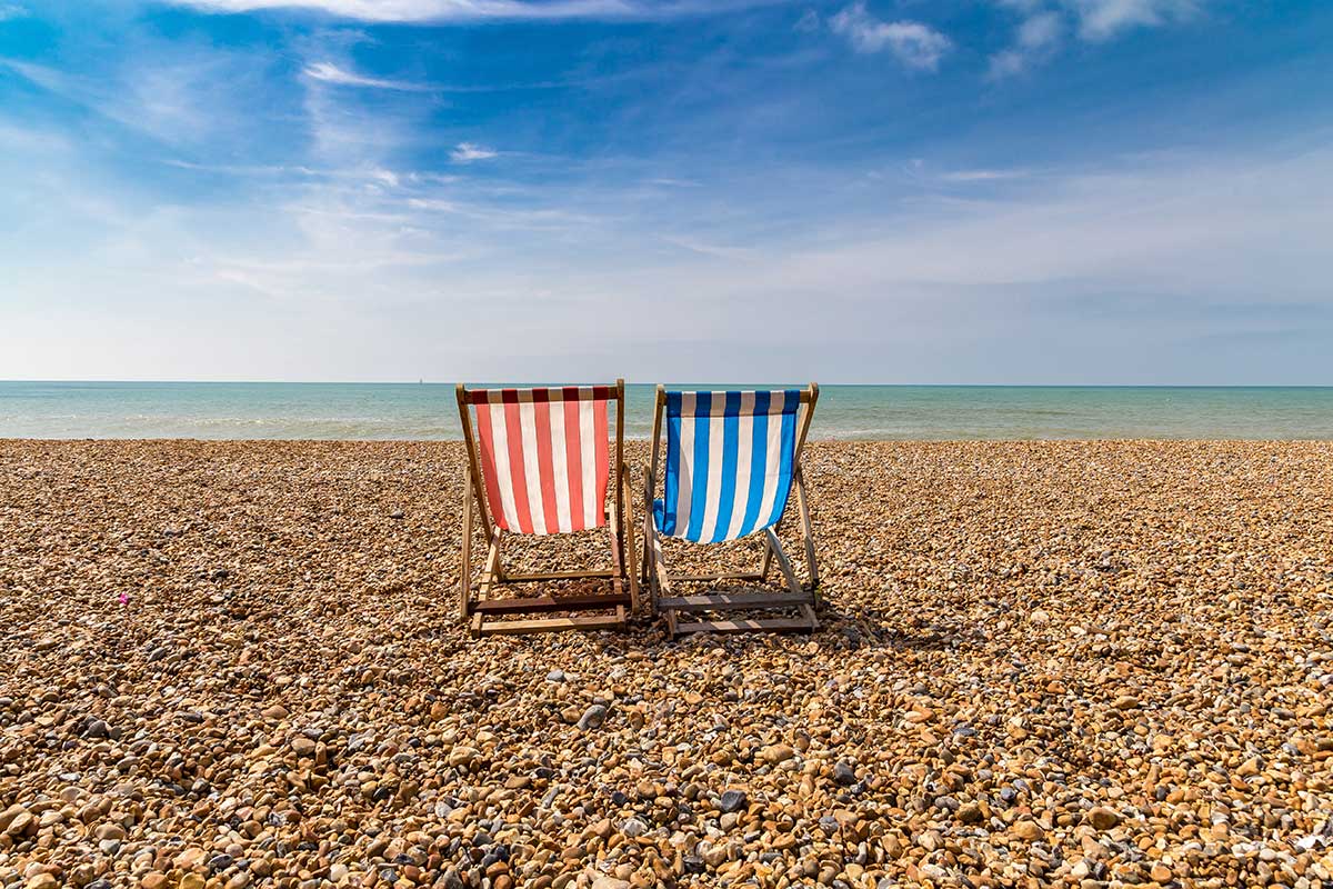 Zwei rote und blaue bunte Liegestühle am steinigen Strand von Brighton bei blauem Meer und blauem Himmel.