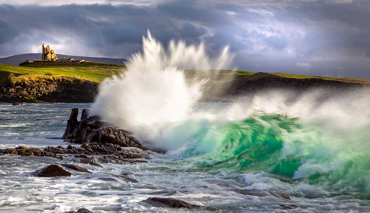 A wave crashing at Mullaghmore beach with a castle in the background in County Sligo, Ireland.