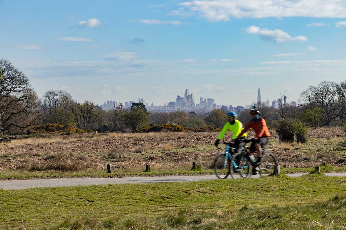 Two cyclists ride their bikes through Richmond Park past luscious greenery and a backdrop of the city of London.