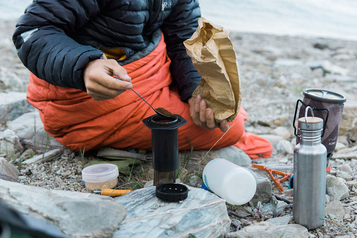 Man camping outdoors in a sleeping bag making coffee using AeroPress