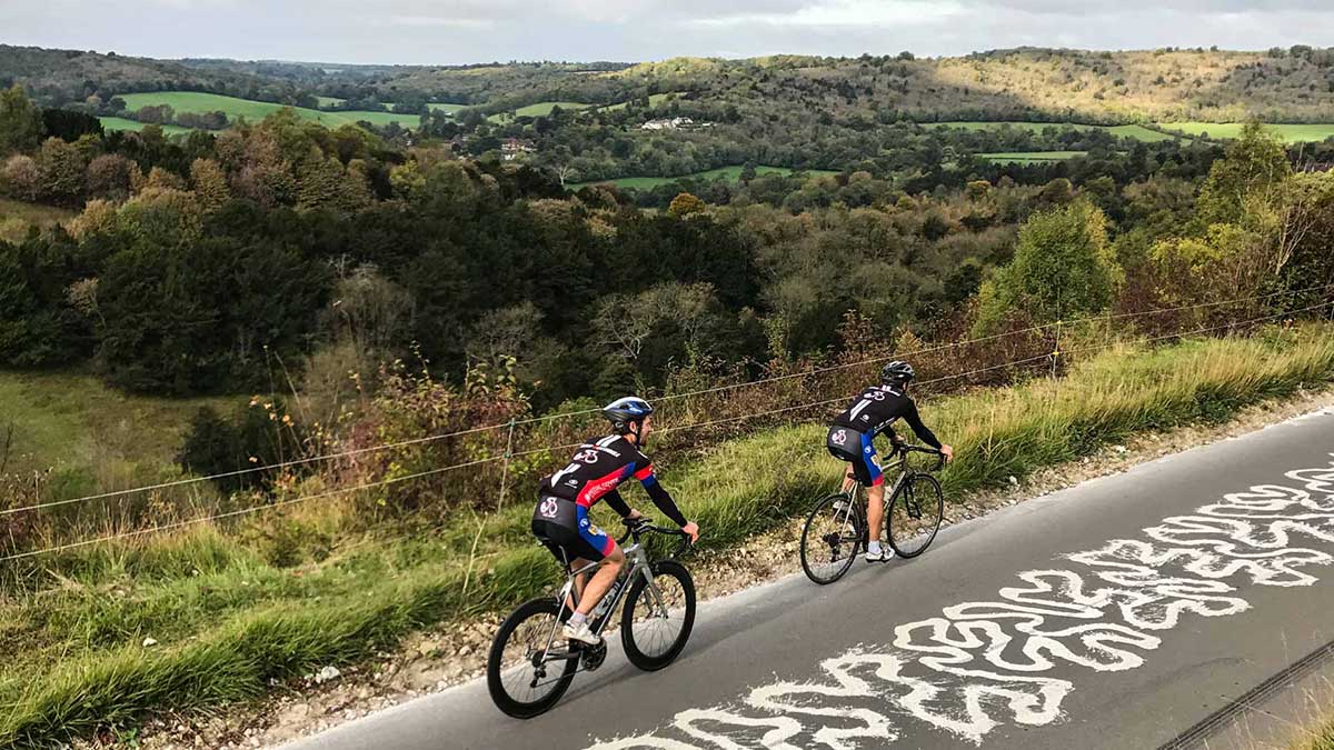 Two cyclists ride road bikes in lycra up Box Hill in Surrey with rolling hills, luscious greenery and beautiful countryside in the background.