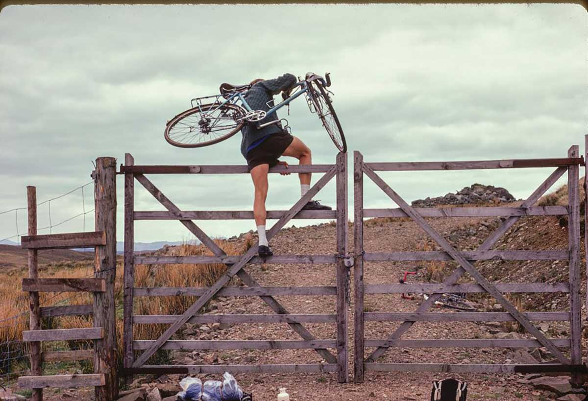 A Rough Stuff Fellowship member carries their bike over a fence on a cycling trip in the countryside.