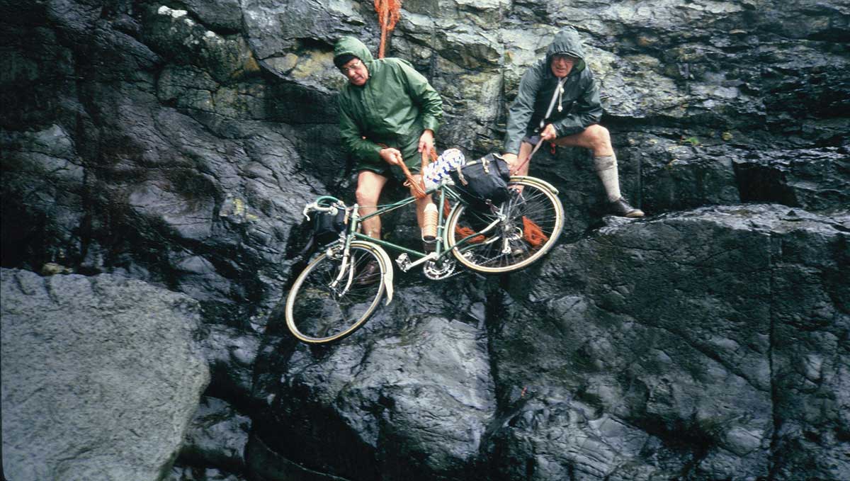 A pair of Rough Stuff Fellowship members on a cycling trip in the countryside carrying their bikes across rocky cliff edges.