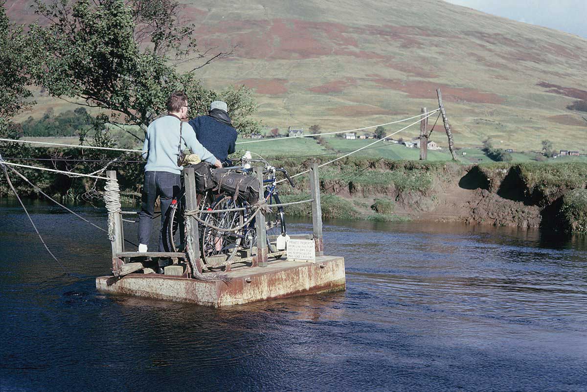 A pair of Rough Stuff Fellowship members on a cycling trip in the countryside taking their bikes across a river on a small floating platform.