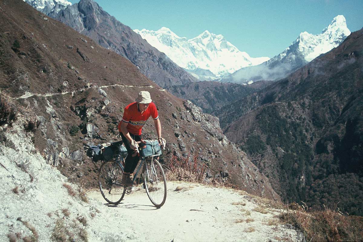 A Rough Stuff Fellowship member with their bicycle on a cycling trip in the mountains with snow-capped peaks, rocky paths and sunshine.