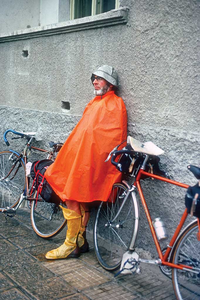 A Rough Stuff Fellowship member rests with their bicycle on a cycling trip in the countryside dressed in a plastic waterproof poncho.