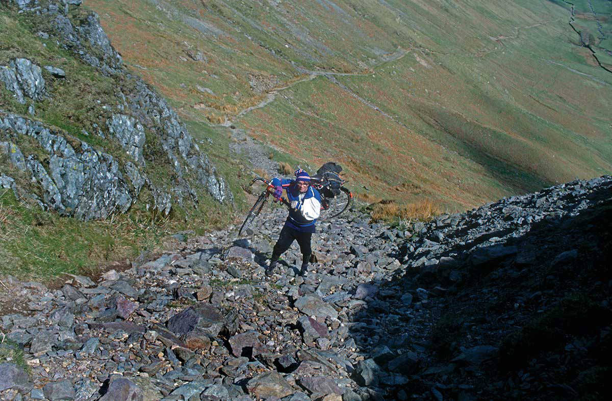 A Rough Stuff Fellowship member on a cycling trip in the countryside carrying their bike over their shoulder up a steep and rocky ascent.