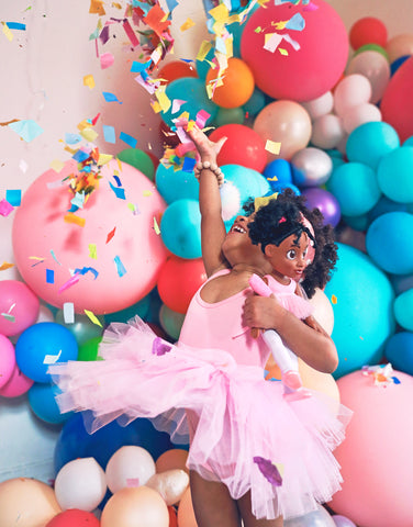 Image of brown ballerina holding a black ballerina doll and throwing confetti in the air.