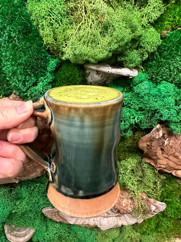 hand holding pottery cup with green foamy drink, with a mossy background with some mushrooms