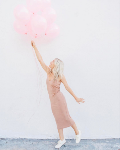 Katie Dean in pink dress and white shoes holding bunch of pink balloons.