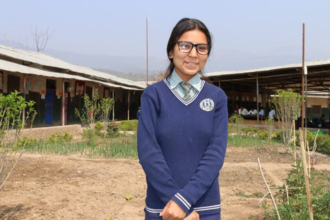Girl wearing school uniform and glasses standing in front of classroom.