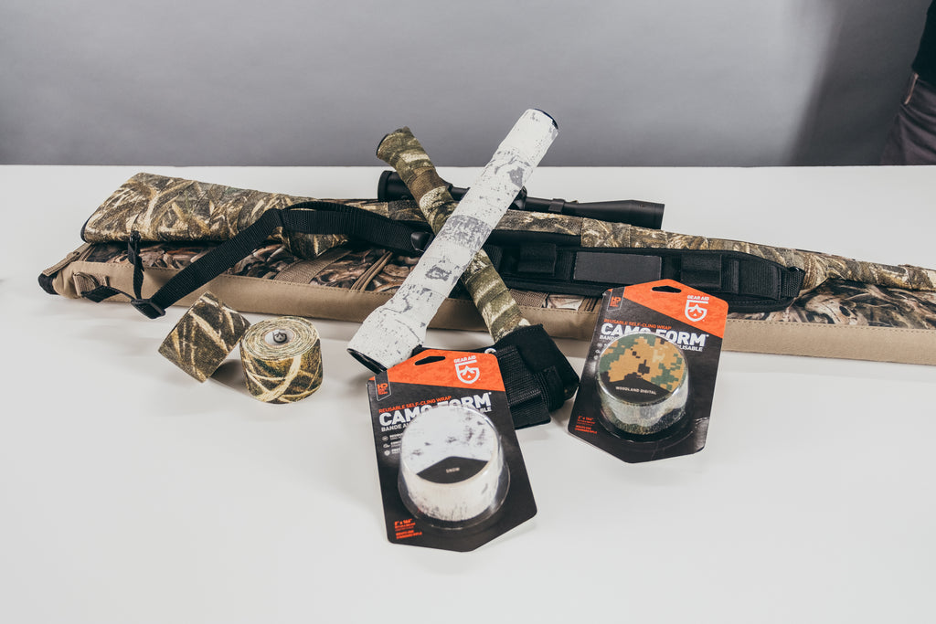 GEAR AID Camo Form products in package sitting against a flashlight and hatchet wrapped in it resting on a camo bag on a white background