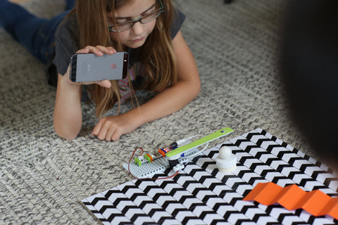 Girl on carpet holding phone over her littleBits invention.