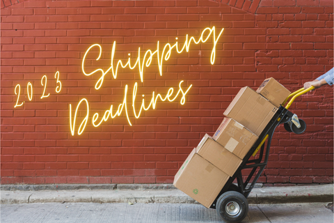 On the top left side of image is slanted text saying 2023 Shipping Deadlines in a Neon glow style yellow; on the right hand side is a stack of boxes on a dolly. Everything is against a brick wall with grey pavement at bottom.