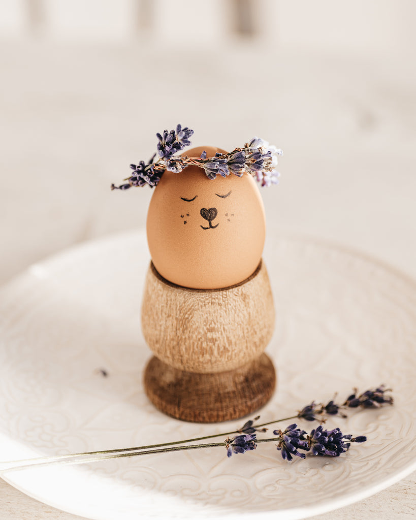 A brown egg with a hand drawn face which is wearing a floral crown of lavendar.