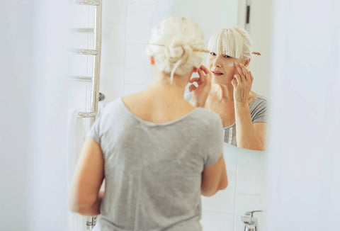 Beautiful senior woman looking in the mirror applying Alastin skincare product