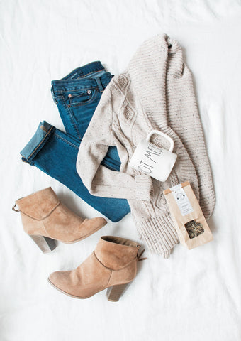 A flat lay photo of a woman's outfit of the day. The outfit consists of a cream colored cardigan, a neutral colored t-shirt, a pair of blue jeans, and ankle boots that she has displayed on top of her bed.