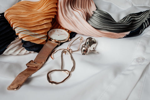 This image shows a stack of various accessories on top of someone's bed. There is a watch, a few bracelets, a hair scarf, and various other accessories displayed.