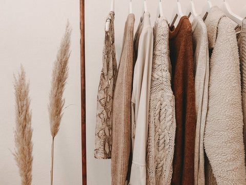 A clothing rack with several neutral colored tops hanging on it. This clothing rack is next to pampas grass and the overall color scheme of the photo is minimalistic and neutral. 