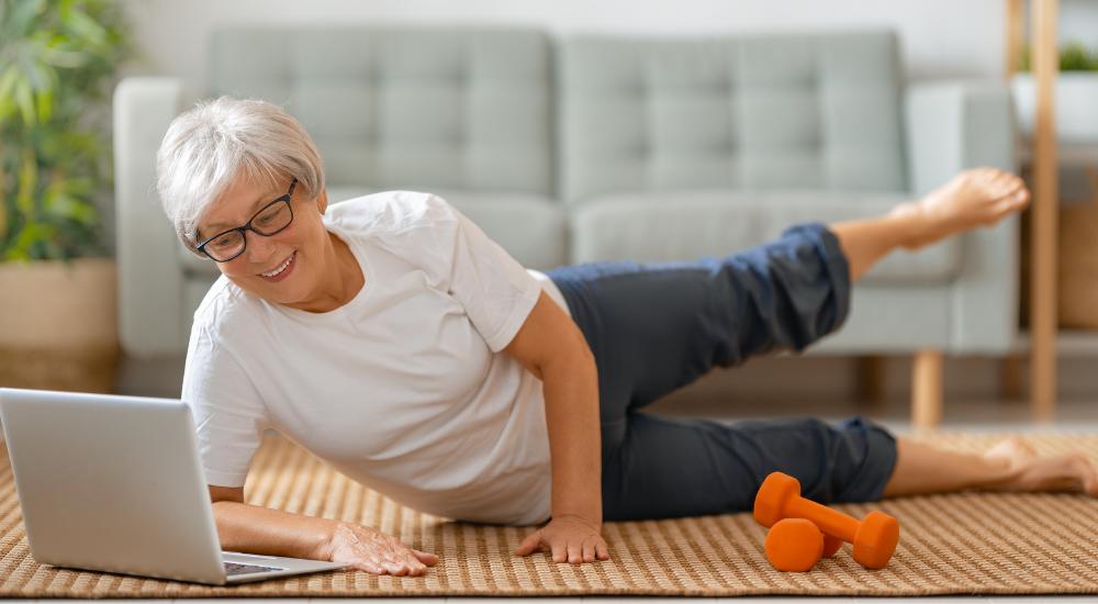 Senior woman on living room floor stretching leg while watching balance exercises on laptop