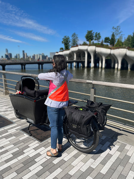 @cargobikemomma in color block reflective hi vis Vespert Eco safety vest from behind with urban arrow family cargo bike by water in NYC