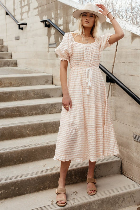 Model wears the Livie Midi Dress in Pink with tan sandals and a beige hat. Dress has tiered skirt, puff sleeves, and tie waist.
