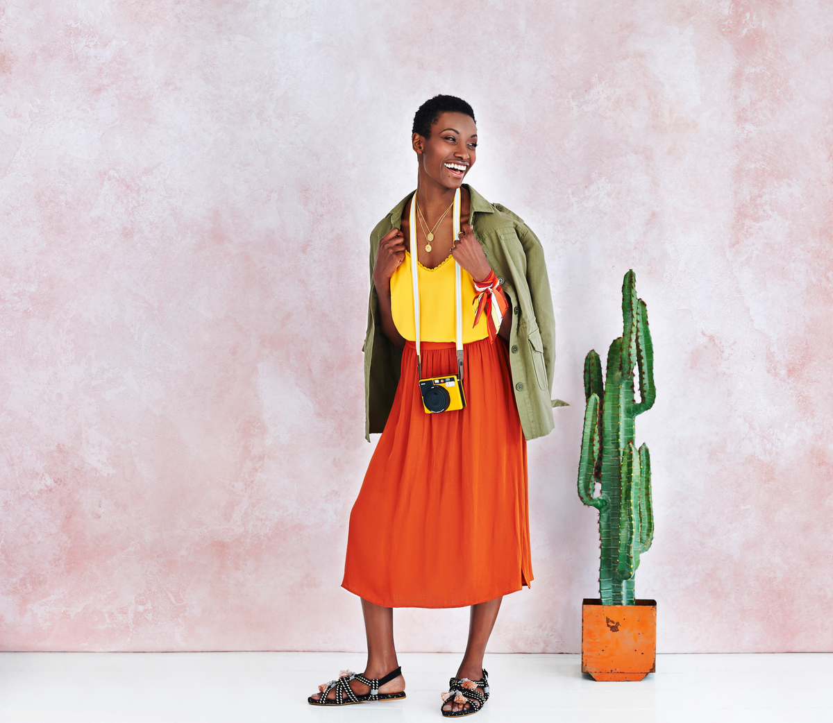 A lady laughing wearing an orange skirt and yellow top with a green khaki shirt over her shoulders and a camera hanging around her neck standing in front of a fabric backdrop printed in a light pink texture with a cactus in a orange metal pot.