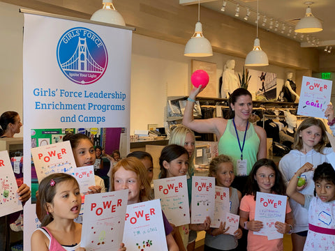 Girls' Force founder Hilary surrounded by young girls holding 'Power' signs, symbolizing women's empowerment and unity