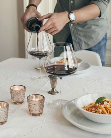 A man is pouring red wine into a Zalto Burgundy glass at a table set for two for dinner.