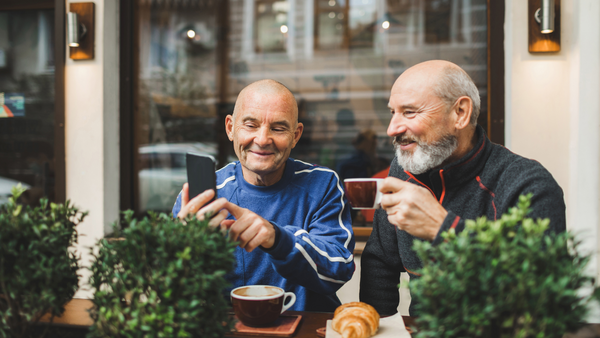 Two older men at an outdoor restaurant. One is holding a cup of coffee, and they are smiling at something on the smartphone that the second man is holding up.