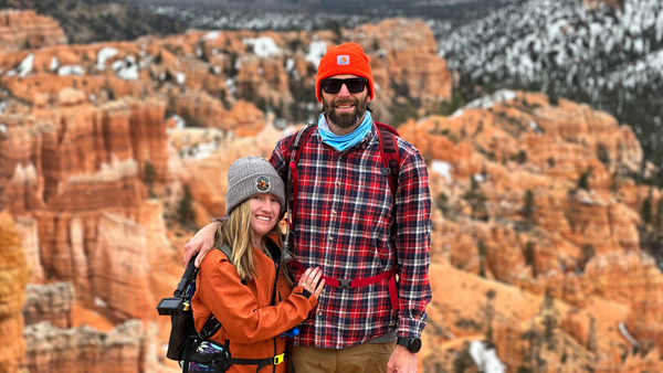 Sunny and Billy hiking in outdoor gear. They’re on a mountain vista, with red stone cliffs behind them, and an obviously huge difference in their heights.