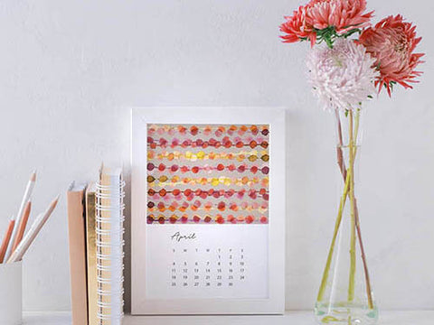 Image shows the April page of Jenny Bova's desk calendar. The design has watercolor circles in rows in reds, yellows, and oranges on a neutral background. The date block is below the image. The calendar is in a white frame on a desk with flowers in a vase and notebooks, pencils and a white wall. 