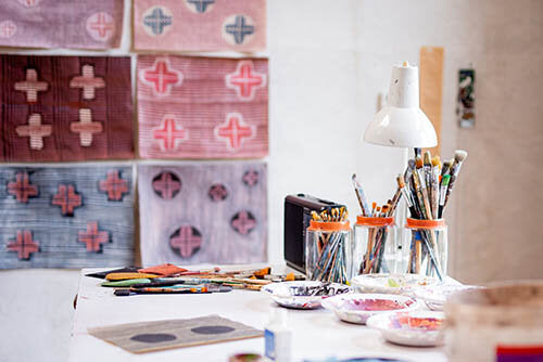 image of an artist's desk. On the wall is pink, blue, purple and white artwork. On the desk is a task lamp, artwork, and various art supplies