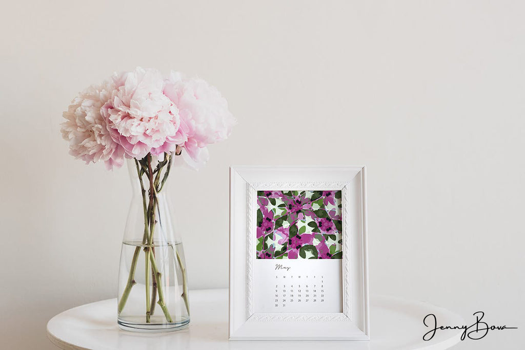 Image of Jenny Bova's May Desk Calendar page in a white frame on a white table. Next to the frame is a vase of pink flowers. The pattern design for the calendar page is an abstract floral in pinks, greens, and whites. 