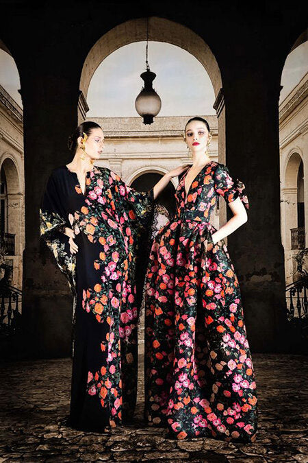 Naeem Kahn image from 2020 fashion shows showing two women in outdoor vestibule wearing long black floral print dresses with bright red, pink, and orange flowers 