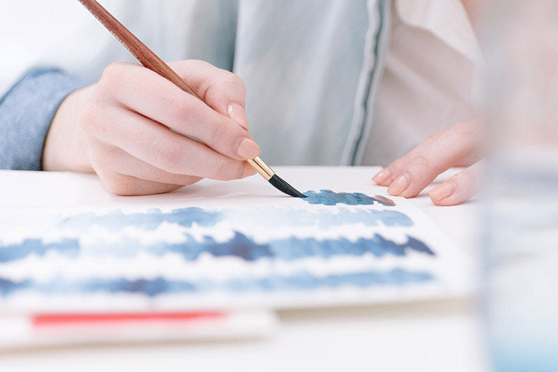 close up image of a person in a light blue denim shirt painting blue watercolor stripes on white paper.  