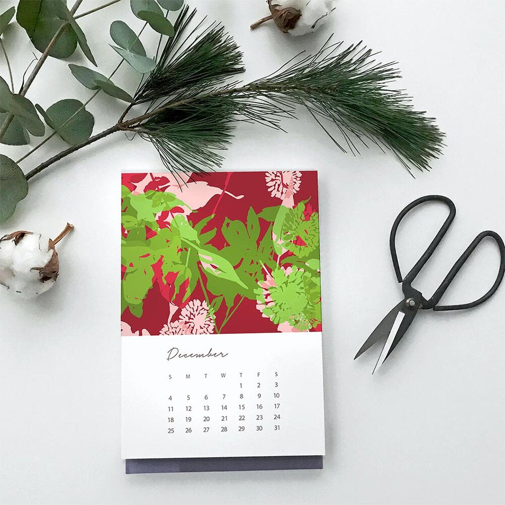 image of Jenny Bova's 2022 Calendar page from December showing red and green leaves and flowers. Next to the calendar page are scissors and evergreen twigs. 