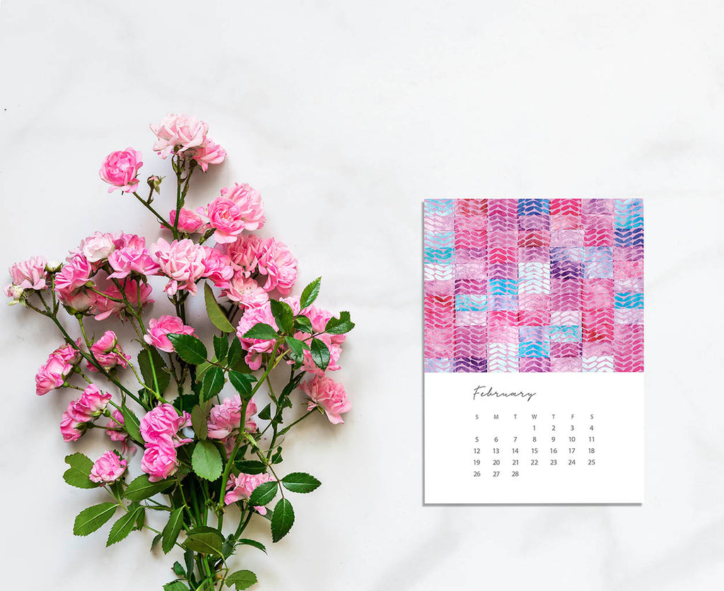 Image of the February page of Jenny Bova's 2023 Desk Calendar. The 5x7 page shows a pink, purple and blue abstract pattern and the days and dates for February 2023 below. Next to the calendar page is a handful of fresh pink flowers and greens. 