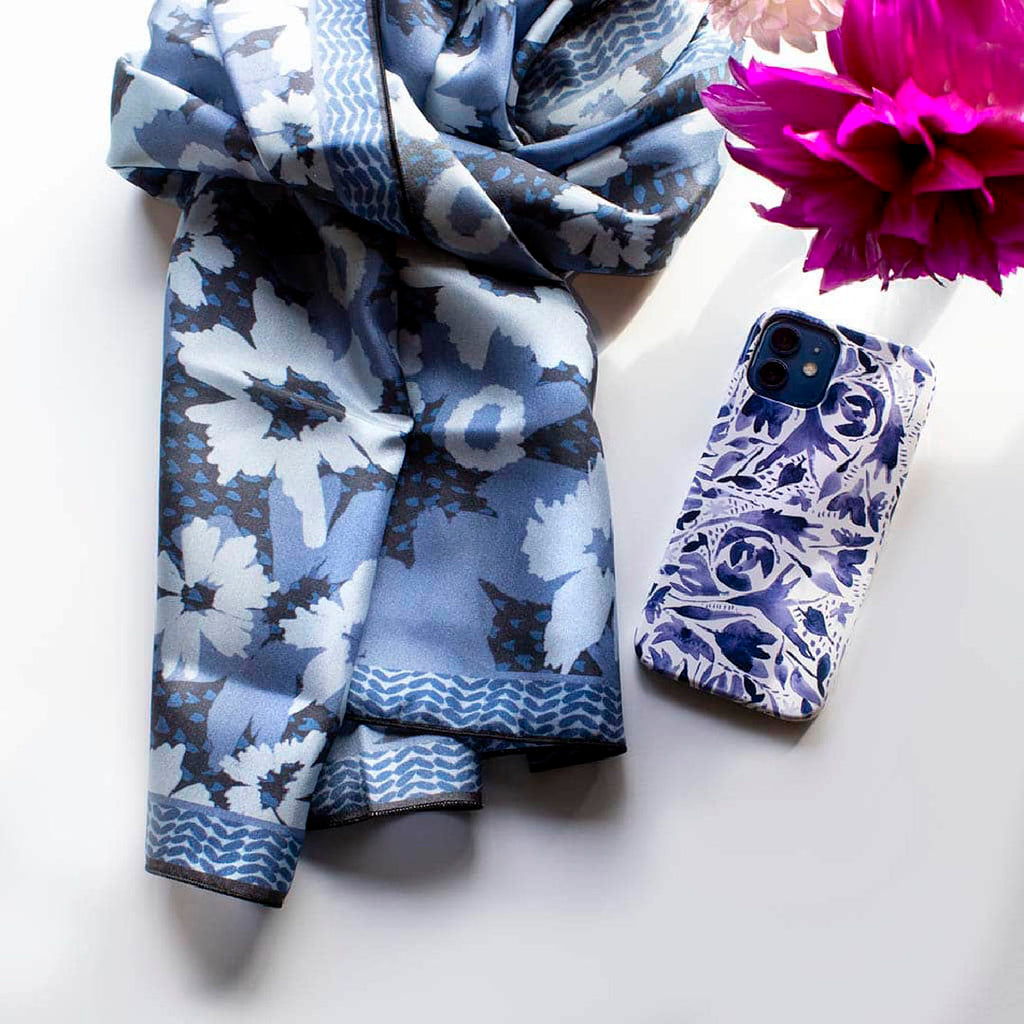 scarf with abstract blue and white pattern and border design folded next to a blue and white iPhone case. In the top right corner are pink flowers