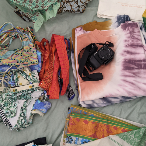 Swimsuits and a tie dye sweatshirt folded on a bed with a camera