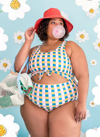 Photo of a woman blowing a bubble and carrying a tote while wearing a multi colored checkered one piece swim suit and a red sun hat