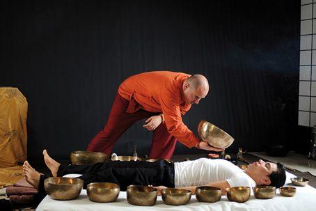 man lying down surrounded by many singing bowls another man holding one bowl nearby
