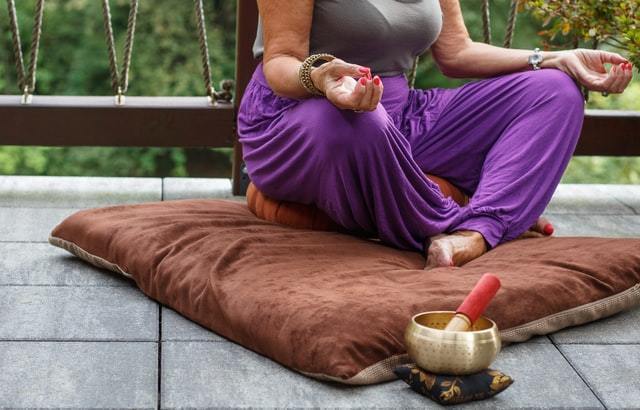 person wearing purple pants sitting in a lotus position on a brown pillow singing bowl and mallet nearby