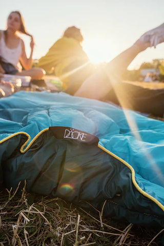 Picknickdecke in blau und grün liegt auf einer Wiese. Darauf sitzt und liegt eine Gruppe von Freunden.