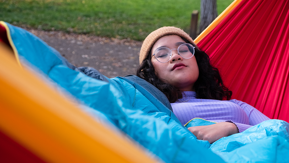 Eine Frau liegt in einer roten Haengematte aus Fallschirmseide unter einer blauen Campingdecke und doest.